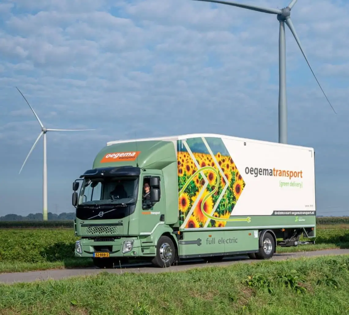 Oegema Transport truck geladen met duurzame, lokaal opgewekte energie, rijdend door een groen landschap met windturbines op de achtergrond.